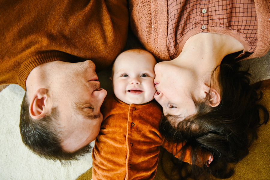 Un bébé souriant allongé entre ses parents, vêtus de tons chauds, lors d'une séance photo de famille à la maison.